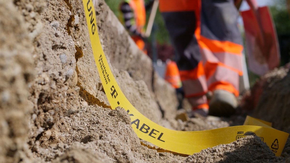 Nahaufnahme eines gelben Trassenwarnbands im Erdaushub einer Baustelle. Im Hintergrund arbeiten Bauarbeiter in orangefarbener Warnschutzkleidung an einem freigelegten Kabel- oder Leitungsschacht.
