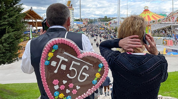 Zwei Personen blicken von einem erhöhten Standpunkt über das  Oktoberfestgelände. Im Vordergrund trägt eine Person eine große Lebkuchenherz-Rückenverzierung mit der Aufschrift „T.56“; dahinter sind Fahrgeschäfte, Buden und zahlreiche Besucher zu sehen.